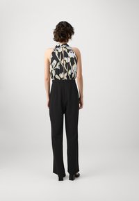 Baksidevisning av en jumpsuit med en ermeløs topp med blomsterprint i svart, hvitt og gult, kombinert med ensfargede, svarte vidde bukser.