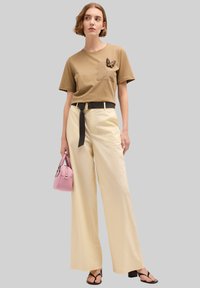Camiseta marrón de manga corta con un parche de mariposa, combinada con pantalones anchos de talle alto en color crema y sandalias de tacón negras. Bolso rosa.