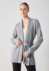 Grå stickad cardigan med ribbad struktur, öppen front, lång ärm och avtagbart knytbälte. Har två framfickor för förvaring.