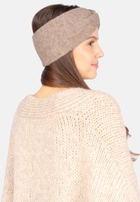 Bandeau tricoté marron avec un design torsadé, fabriqué en matériau doux. Porté par un modèle aux cheveux longs, assorti avec un pull beige clair.