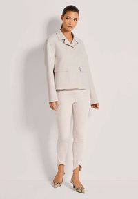 Frau in einer beigen Jacke mit großen Taschen, hellbeigen Jeans mit fransigem Saum und gemusterten High Heels steht vor einer schlichten Wand.