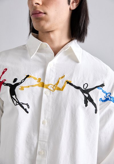 White button-up shirt featuring colorful embroidered figures in black, yellow, red, and blue. Soft cotton fabric with a classic collar.