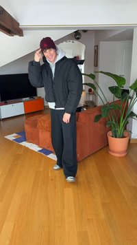 Sort polstret jakke med grå hettegenser under, svarte joggebukser og sølvsko. Burgunder lue. Interiør med tregulv og planter.