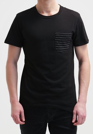 Mann trägt ein schwarzes Kurzarm-T-Shirt mit einer schwarzen Tasche auf der linken Brust, verziert mit kleinen weißen Nieten.