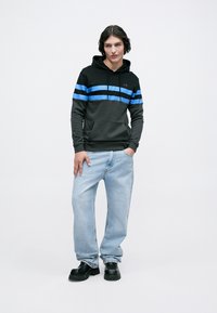 Jovem com um hoodie preto com riscas azuis, jeans largos de tom azul claro e sapatos pretos robustos, de pé com uma mão no bolso.