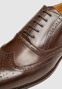 Scarpa da abito in pelle marrone con design a punta d'ala, dettagli brogue e sei occhielli. Texture liscia con finitura lucida e cuciture a contrasto.