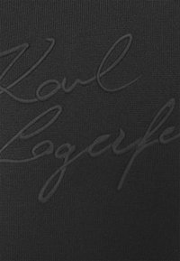 Svart stickad tyg med en upphöjd signaturdesign i en mörkare nyans av svart, vilket skapar en subtil textur över ytan.