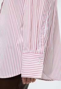 Λευκό πουκάμισο με μακριά μανίκια, λεπτές κάθετες κόκκινες ρίγες και δύο λευκά κουμπιά στο μανίκι, φορεμένο με σκούρο παντελόνι.