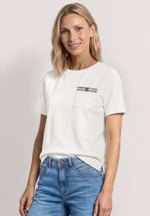Femme aux cheveux blonds portant une chemise blanche à manches courtes avec une petite poche poitrine à motifs et un jean bleu, debout devant un fond uni.