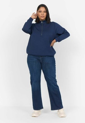 Marineblauwe half-zip pullover van zachte stof, gecombineerd met flared blauwe jeans en lichte sneakers, staand tegen een witte achtergrond.