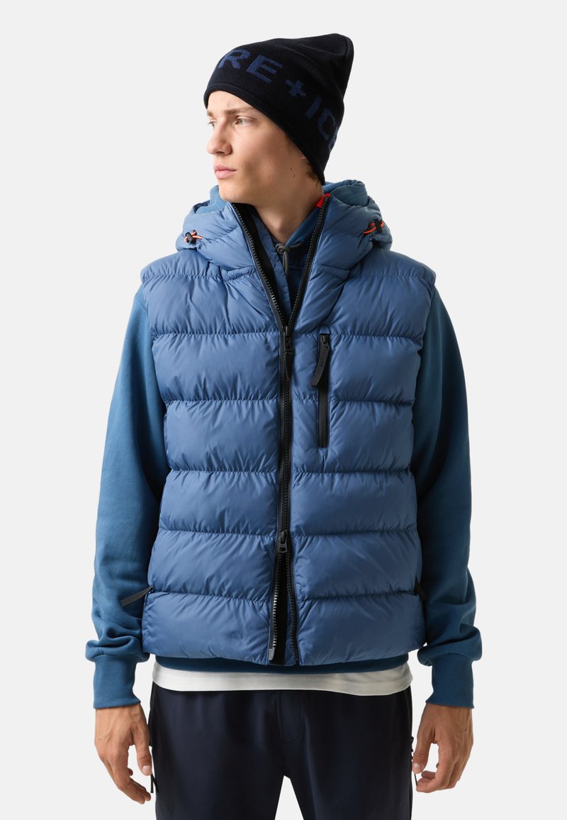 Blaue gefütterte Weste mit Kapuze, mit gestepptem Design, Reißverschluss und schwarzen Akzenten. Getragen über einem blauen Sweatshirt und navyfarbener Hose.