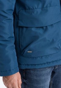 Modrá zateplená bunda s velkou přední kapsou, hladkým povrchem a decentním logem na lemu. Má vyšívané švy a rovný střih.