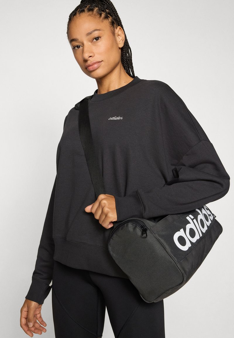 Sweat-shirt de sport noir avec poignets côtelés et logo brodé, accompagné d'un sac noir avec un logo blanc et une fermeture éclair.