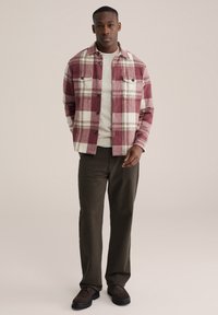 Rot-weiß karierte Flanellbluse über einem beigen Pullover, kombiniert mit dunkelgrünen Hosen und braunen Schuhen. Zwei Brusttaschen an der Bluse.