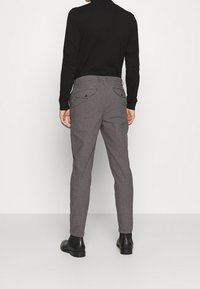 Man som bär grå slim-fit byxor, svart polotröja och svarta läderstövlar vid ankeln, stående mot en slät vit bakgrund.