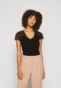 Top negro de canalé con mangas cortas de encaje y escote con forma de ola, combinado con pantalones beige de cintura alta y un collar dorado sencillo.
