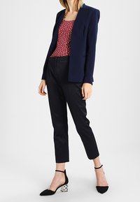 Femme portant un blazer bleu marine, un top à motifs rouges, un pantalon noir texturé à la cheville et des talons noirs avec des blocs décoratifs.