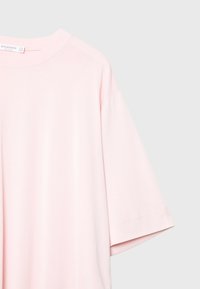 Světlé růžové oversized tričko s kulatým výstřihem, krátkými rukávy a hladkým bavlněným textilním materiálem. Má jednoduchý design bez vzorů.
