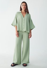 Camisa de algodón texturizada en verde claro, con cuello y botones, combinada con pantalones anchos a juego y sandalias negras.