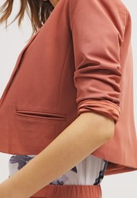 Koralowa krótka marynarka o gładkiej fakturze, z rękawami do połowy przedramienia i jedną przednią kieszenią. Charakteryzuje się dopasowanym, taliowanym krojem.