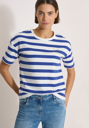 Mujer con el cabello corto y castaño, que lleva una camiseta blanca con rayas azules y vaqueros azules, de pie con las manos detrás de la espalda sobre un fondo neutral.