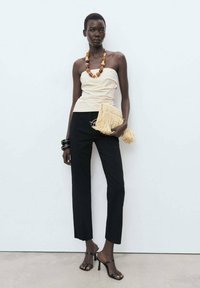 Haut beige sans manches, pantalon noir cintré, talons noirs à lanières et une pochette beige à franges. Les accessoires incluent un collier en perles multicolores.