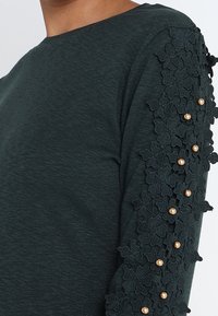 Top vert foncé avec un col rond. Présente des appliqués en dentelle sur les manches avec des perles de couleur dorée. Texture douce et coupe ajustée.
