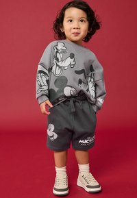 Harmaa huppari, jossa on mustia Mickey Mouse ja ystävät -grafiikoita, yhdistettynä samanvärisiin harmaisiin shortseihin, joissa on logo. Lapsella on päällä vaaleanväriset lenkkarit.