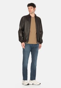 Giacca bomber in pelle marrone con colletto a coste nero; abbinata a una camicia beige e jeans blu, indossata con sneaker bianche.
