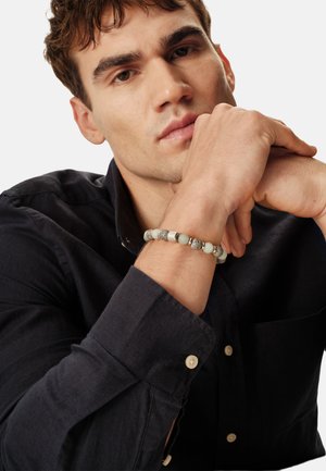 Brazalete de cuero con cuentas de metal plateado y piedras blancas lisas, llevado en una muñeca, con una camisa oscura abotonada en el fondo.