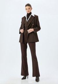 Brauner maßgeschneiderter Anzug mit einem Doppelreiher-Blazer, einem weißen Kragenhemd darunter und weit geschnittenen Hosen. Schwarze High-Heels-Stiefel.