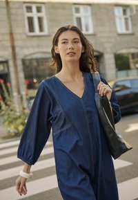 Denim kombinezon z V-izrezom, dolgimi napihnjenimi rokavi in kontrastnim šivanjem. Dopolnjen z črno torbo cez ramo in teksturiranim zapestnim manšetom.