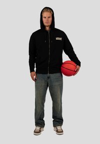 Černá mikina s kapucí na zip a logem, modré denimové džíny a hnědé boty. Držící jasně červený basketbal, stojící na šedém pozadí.