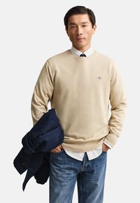 Homme portant un pull beige par-dessus une chemise blanche et une cravate, tenant une veste bleu foncé dans une main, l'autre main dans la poche du jean.