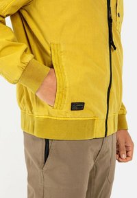 Gelbe Jacke mit gerippten Bündchen und Saum, mit einem Reißverschluss vorne und seitlichen Taschen. Enthält ein Textiletikett in der Nähe des Saums.