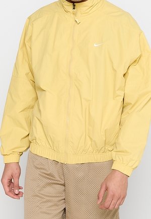 Mężczyzna ubrany w bladoróżową żółtą kurtkę na zamek firmy Nike z elastycznymi mankietami i ściągaczem na dole, w połączeniu z beżowymi perforowanymi szortami.