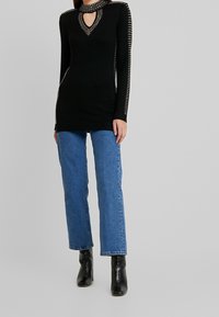 Top negro de manga larga con escote de ojo de cerradura, detalles de tachuelas a lo largo de los hombros y un diseño ajustado. Combinado con jeans rectos azules.