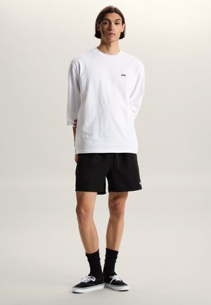 Młody mężczyzna stojący, ubrany w białą, długorękawową koszulkę marki Vans, czarne szorty, czarne skarpetki oraz czarne sneakersy Vans, na jasnym tle.