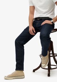 Jeans de denim azul oscuro con un corte slim, costuras contrastantes y diseño de cinco bolsillos. Llevados con una camisa blanca y zapatillas altas beige.