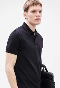 Melna polo krekls ar formētu apkakli, īsām piedurknēm un nelielu logotipu uz krūtīm. Audums ar fakturētu virsmu.