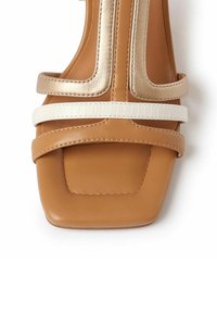 Tanfarvet blokhælsandal med metallic guld- og hvide stropper. Firkantet tådesign med en glat lædertekstur og kontrasterende accenter.