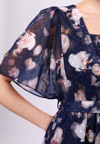 Marinblå klänning med blommönster, genomskinliga puffärmar, strukturerade prickar och knytmidja. Har vita och rosa blommönster.