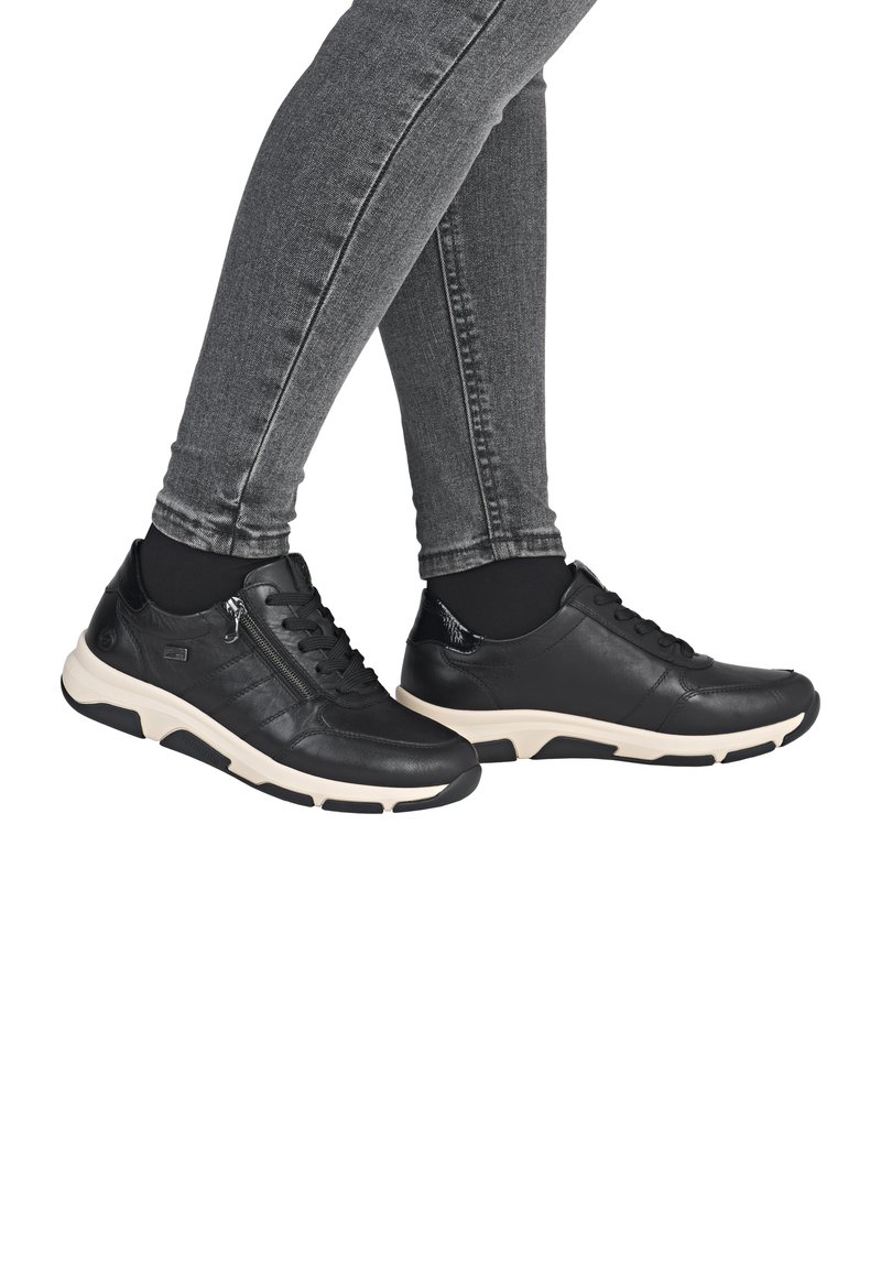 Schwarze Leder-Sneaker mit einer cremefarbenen Sohle, ausgestattet mit Schnürsenkeln, einem seitlichen Reißverschluss und strukturierten Akzenten, präsentiert auf grauen Jeans.