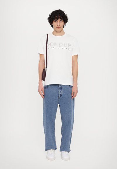 T-shirt en coton blanc avec du texte gris imprimé, associée à un jean en denim bleu ample. Le modèle tient un petit sac marron et porte des baskets blanches.
