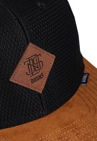 Svart kaps med teksturert stoff på toppen, brun semsket skygge, og en diamantsformet brun skinnlapp med merkevaren sitt logo.