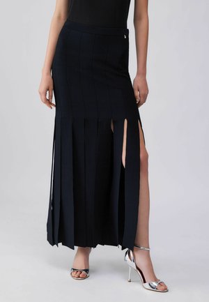 Granatowa długa spódnica z pionowymi plisami i bocznymi rozcięciami, wykonana z teksturowanej tkaniny. Połączona z srebrnymi sandałami na wysokim obcasie dla kontrastu.