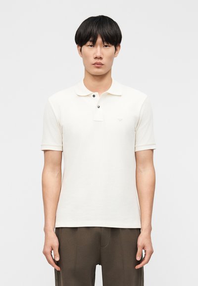 Jeune homme aux cheveux noirs courts portant un polo blanc à manches courtes uni et un pantalon sombre, debout devant un fond uni.
