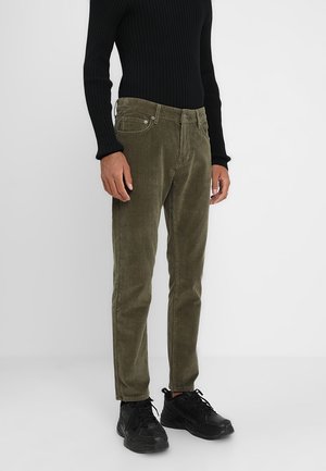 Homme portant un pantalon en velours côtelé vert olive, un haut à manches longues noir côtelé et des baskets noires, debout devant un fond blanc uni.