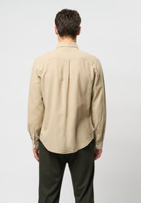 Cămașă bej cu mâneci lungi, confecționată dintr-un material texturat, cu tiv curbat, guler rotund și manșete cu nasturi. Poartă pantaloni verde închis.