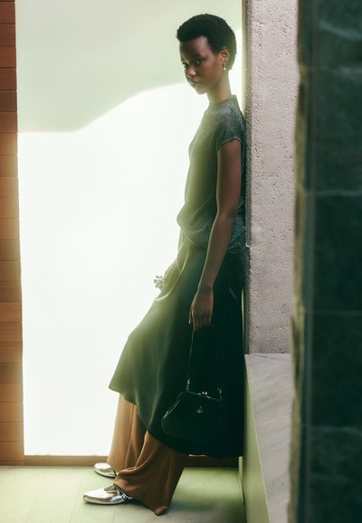 Borsa nera texture, scarpe argentate, top metallico e pantaloni marroni fluidi. La modella si appoggia a un muro con uno sfondo di luce soffusa.